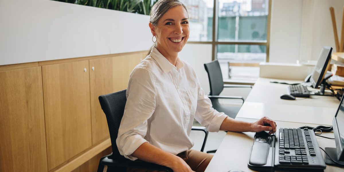 Friendly receptionist smiling at camera while sitting at desk with computer in office. Happy female front desk employee at work.