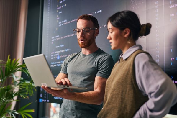 Two young adult programmers are discussing code on a laptop in a modern office, collaborating on a software development project