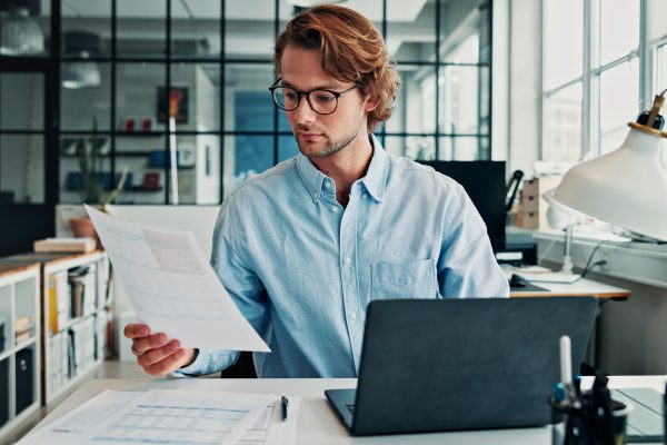 Review, document and man in office with laptop, glasses or preparation of financial statement. Reading, audit paper or analyst in workplace with tech, data entry or balance sheet in budget management