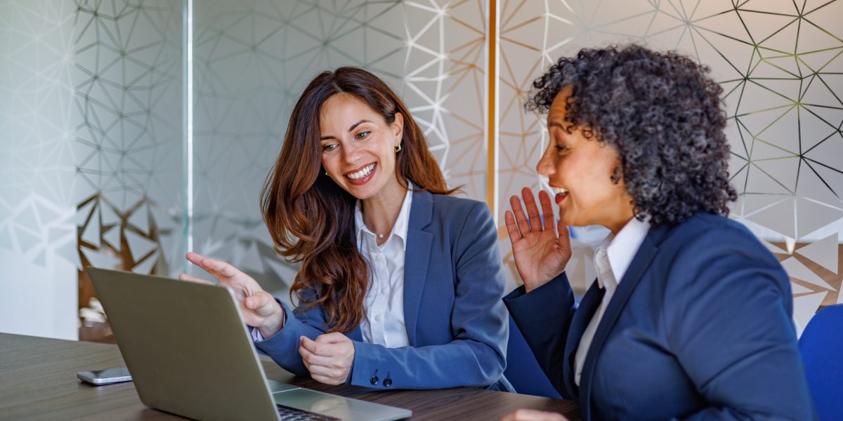 Two businesswomen discussing work on a laptop, smiling and brainstorming in a professional office environment